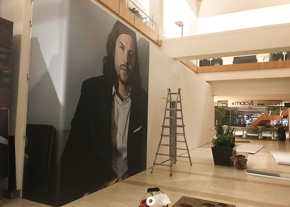 progress of wall wrap installation showing that a menswear retail store in a shopping mall is opening soon behind a ladder